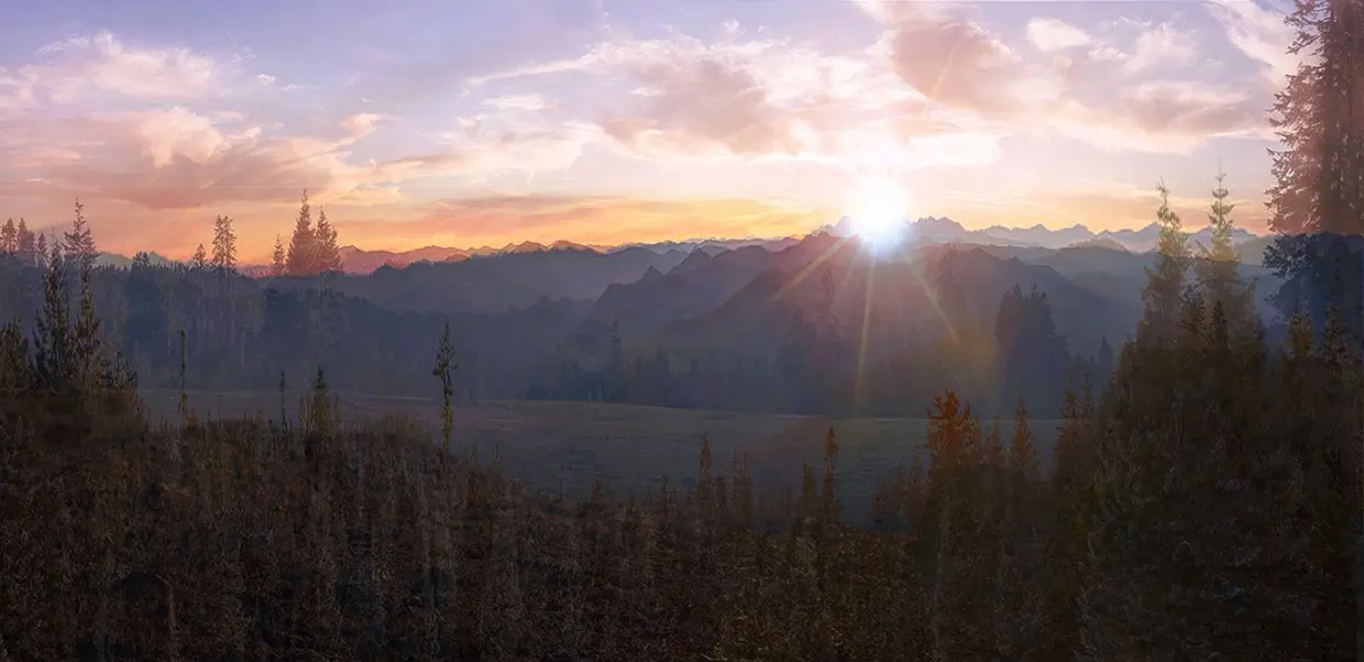 Sunrise over a mountain range with a field and trees in the foreground, and a colorful sky.