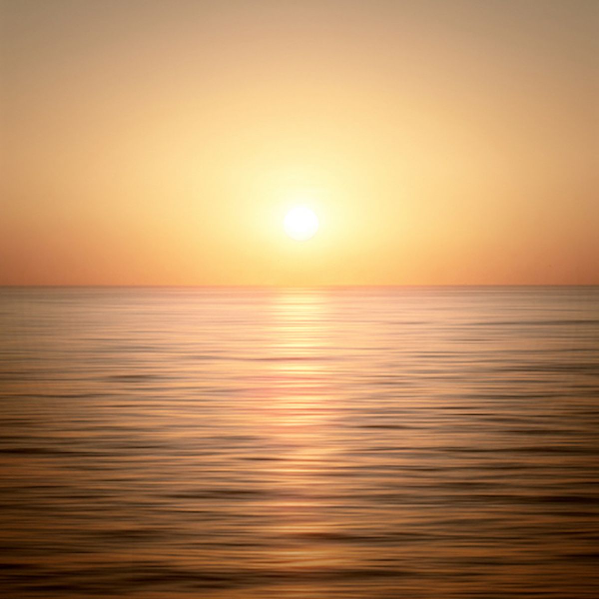 A serene sunset over a calm ocean with the sun low in the sky, casting warm orange and yellow hues across the water.