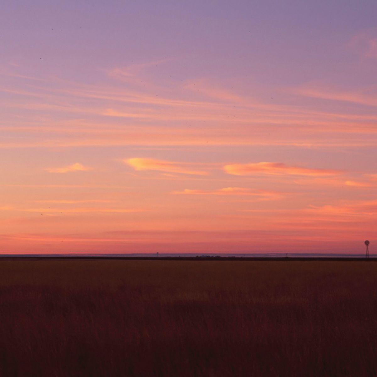 Sunset over a wide field with a silhouette of a distant structure against a colorful sky of pink and orange clouds.